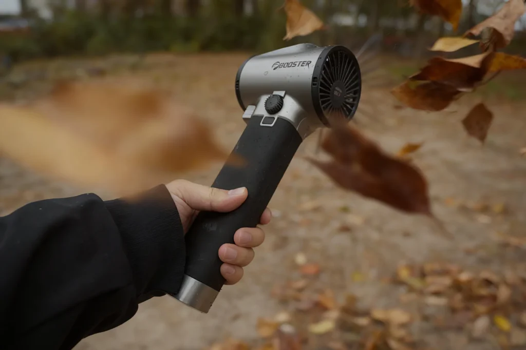 A hand holds a leaf blower vacuum, blowing dry leaves outdoors.